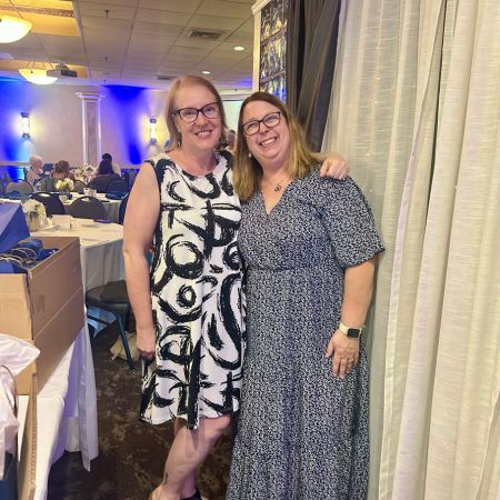 Two smiling co-teachers stand together at an event, wearing patterned dresses, highlighting their friendship and partnership in education.