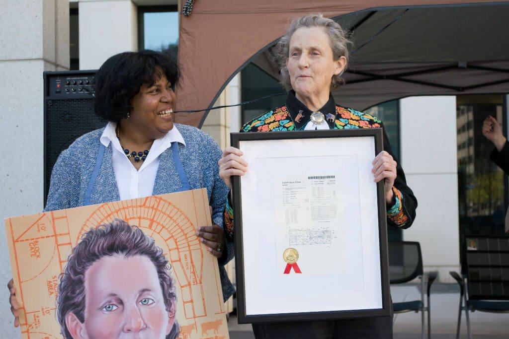 Temple Grandin stands outdoors holding a framed patent document, while another adult stands beside her holding a painted portrait. Both appear at a public event, highlighting Grandin’s contributions to science and innovation.